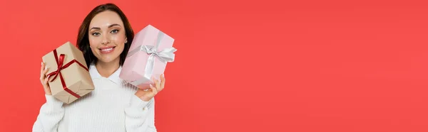 Cheerful young woman in sweater holding presents isolated on red, banner — Stock Photo