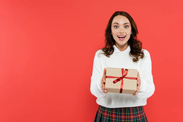 Glückliche junge Frau im Pullover mit Geschenkbox und Blick in die Kamera isoliert auf rot — Stockfoto