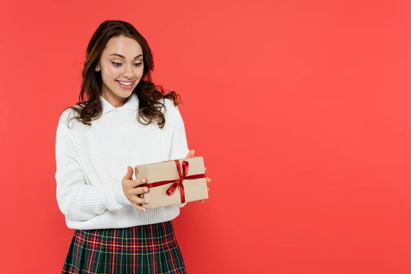 Позитивна молода жінка в теплому светрі дивиться на подарунок ізольовано на червоному — стокове фото