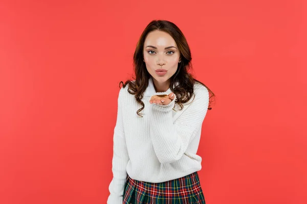 Mujer morena en jersey y falda a cuadros soplando aire beso aislado en rojo - foto de stock