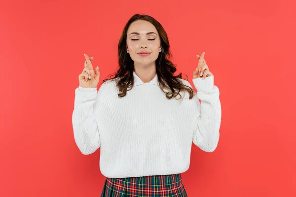 Young woman in white jumper crossing fingers and closing eyes isolated on red — Stock Photo