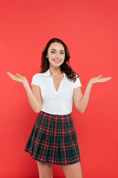 Fröhliche brünette Frau in kariertem Rock und T-Shirt zeigt mit den Händen auf rotem Hintergrund — Stockfoto