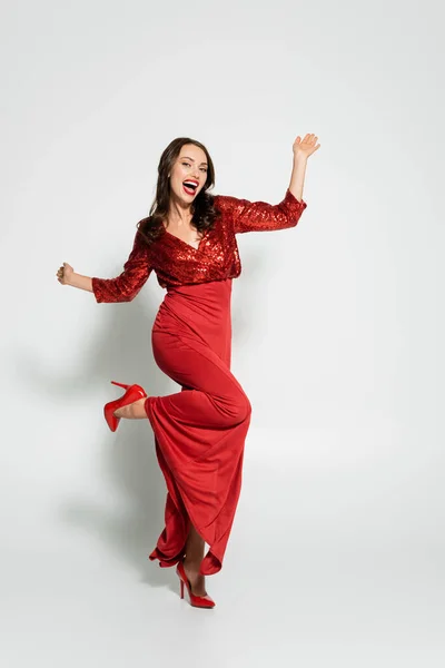 Longitud completa de la mujer con estilo en tacones y vestido rojo mirando a la cámara sobre fondo gris - foto de stock