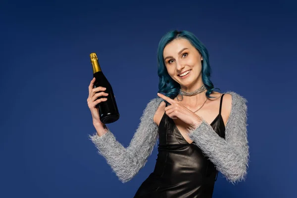 Cheerful woman in black leather dress pointing at champagne bottle isolated on blue — Stock Photo