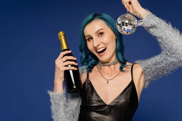 Excited woman with colored hair holding champagne bottle and shiny disco ball isolated on blue — Stock Photo