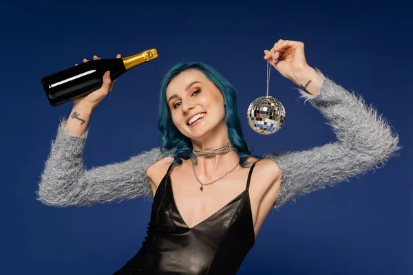 Радісна і модна жінка з блискучою диско-кулькою і пляшкою шампанського ізольована на синьому — стокове фото