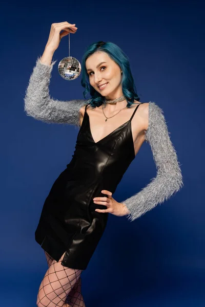 Young woman in black leather dress and faux fur sleeves posing with hand on hip and disco ball isolated on blue — Stock Photo