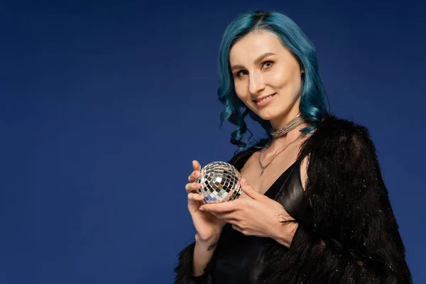 Stylish woman in black faux fur jacket holding small and shiny disco ball isolated on blue — Stock Photo