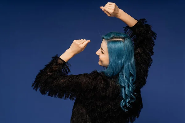 Вид ззаду жінки з кольоровим волоссям і в чорній хутряній куртці танцює під час новорічної вечірки ізольовані на синьому — стокове фото