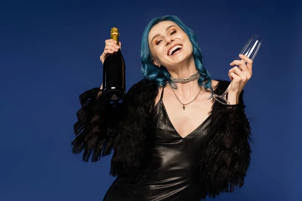 Excited woman in black dress and faux fur jacket holding champagne and laughing isolated on blue — Stock Photo