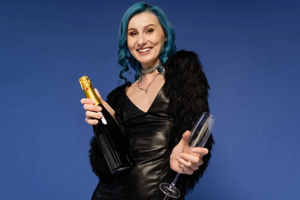 Trendy woman in black faux fur jacket and leather dress holding champagne and smiling at camera isolated on blue — Stock Photo