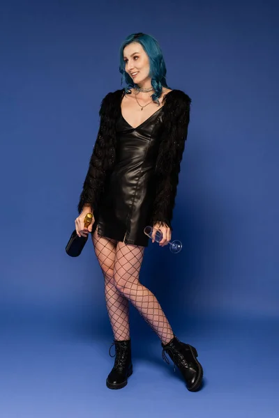 Fashionable woman in black leather jacket and fishnet tights holding champagne on blue background — Stock Photo