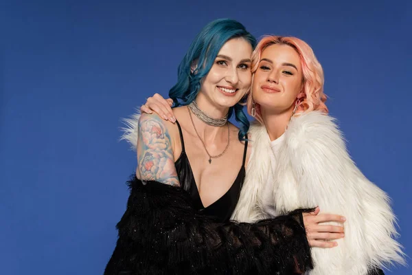 Glückliche queere Freunde in schwarz-weißen Kunstpelzjacken, die isoliert auf blauem Grund in die Kamera lächeln — Stockfoto