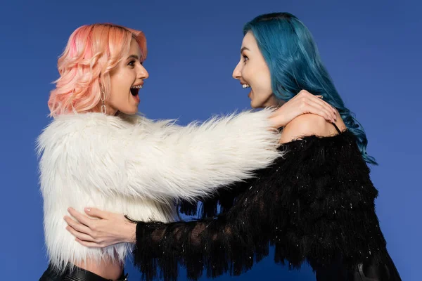 Side view of amazed queer friends in black and white faux fur jackets looking at each other isolated on blue — Stock Photo