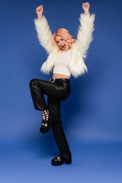 Full length of thrilled woman in black leather pants and white faux fur jacket dancing with raised hands on blue background — Stock Photo