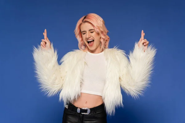 Excited woman in fluffy faux fur jacket holding crossed fingers isolated on blue — Stock Photo