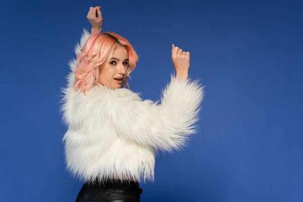 Happy woman in white faux fur jacket dancing and looking at camera isolated on blue — Stock Photo