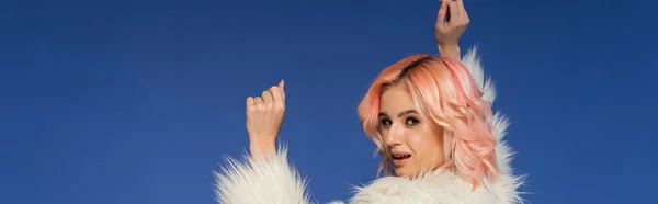 Woman in white and fluffy faux fur jacket looking at camera while dancing isolated on blue, banner — Stock Photo