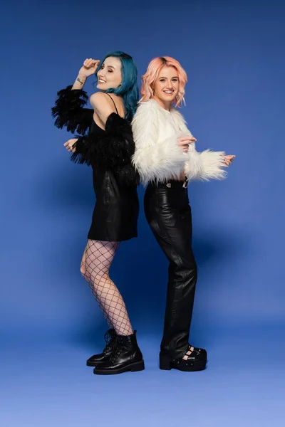 Full length of stylish queer friends in faux fur jackets dancing back to back on blue background — Stock Photo