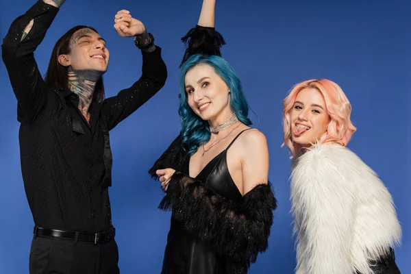 Cheerful queer person sticking out pierced tongue near dancing nonbinary friends isolated on blue — Stock Photo