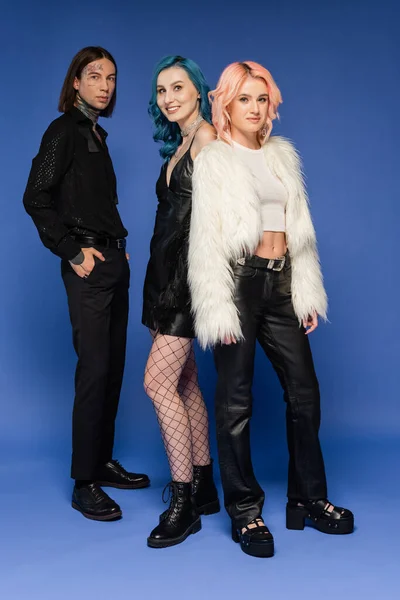 Tattooed nonbinary person standing with hand in pocket near smiling queer friends on blue background — Stock Photo