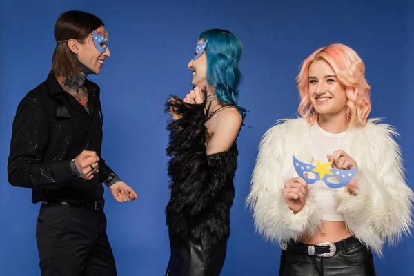 Queer person holding party mask and looking at camera near nonbinary people dancing isolated on blue — Stock Photo