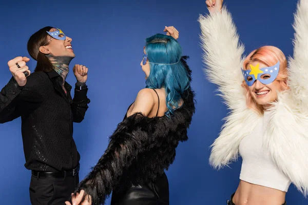 Nonbinary person in white faux fur jacket and party mask dancing with raised hands near queer friends isolated on blue — Stock Photo