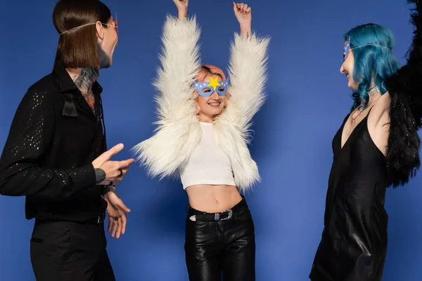 Fröhliche queere Person in weißer Kunstpelzjacke, die mit erhobenen Händen neben nichtbinären Freunden tanzt, isoliert auf blauem Grund — Stockfoto