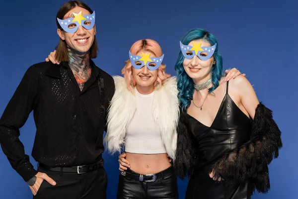 Tattooed nonbinary person standing with hand in pocket near cheerful queer friends in party masks isolated on blue — Stock Photo