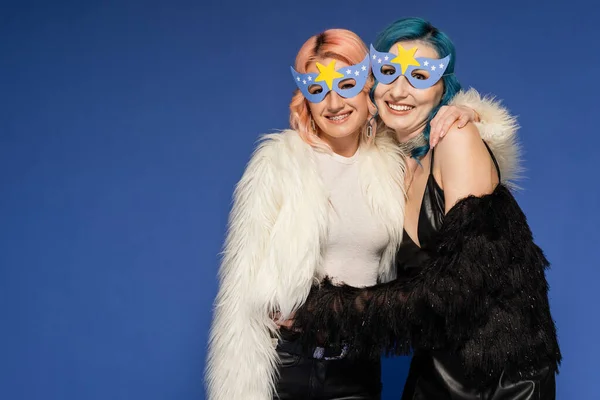 Joyeuses personnes queer dans des masques de fête et des vestes en fausse fourrure embrassant isolé sur bleu — Photo de stock