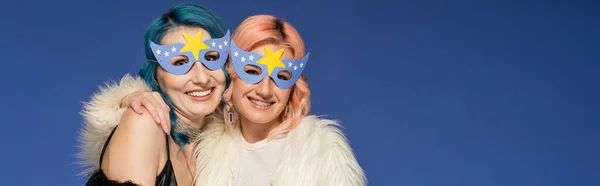 Щасливі небінарні друзі в партійних масках обіймаються ізольовано на синьому, банер — стокове фото