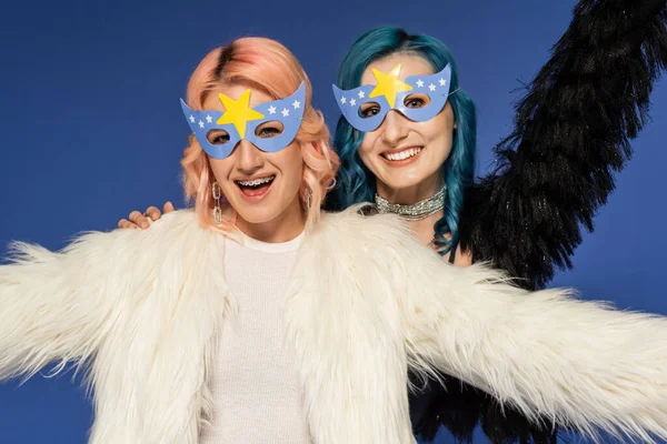 Amigos queer animado em máscaras de festa e casacos de pele falsos macios olhando para a câmera isolada em azul — Fotografia de Stock