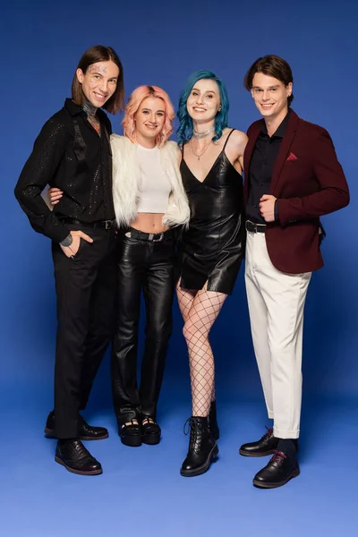 Full length of joyful nonbinary friends in stylish clothes smiling at camera on blue — Stock Photo