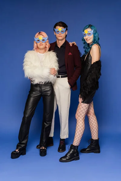 Full length of nonbinary people in fashionable clothes and party masks looking at camera on blue background — Stock Photo