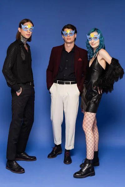 Full length of stylish queer friends in party masks looking at camera while standing on blue background — Stock Photo