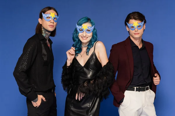Nonbinary friends in party masks standing with hands in pockets near queer person in black leather dress isolated on blue — Stock Photo