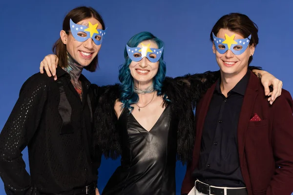 Gay feliz pessoa em vestido de couro preto abraçando amigos não binários em máscaras de festa isolado em azul — Stock Photo