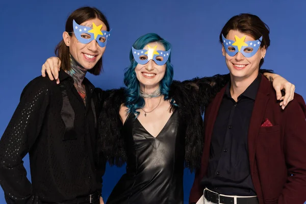 Heureux queer personne en robe en cuir noir embrassant des amis non binaires dans des masques de fête isolé sur bleu — Photo de stock