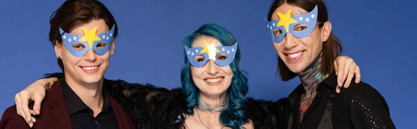 Веселый небинарный человек обнимает голубых друзей в масках для вечеринок и улыбается в камеру, изолированную на голубом, баннер — стоковое фото
