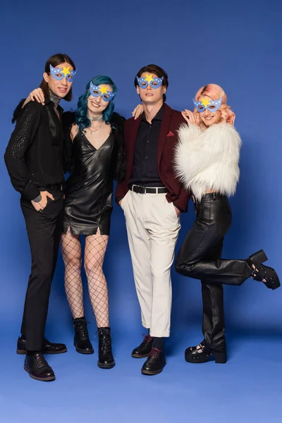 Tattooed queer people standing with hands in pockets near stylish nonbinary friends in party masks on blue — Stock Photo