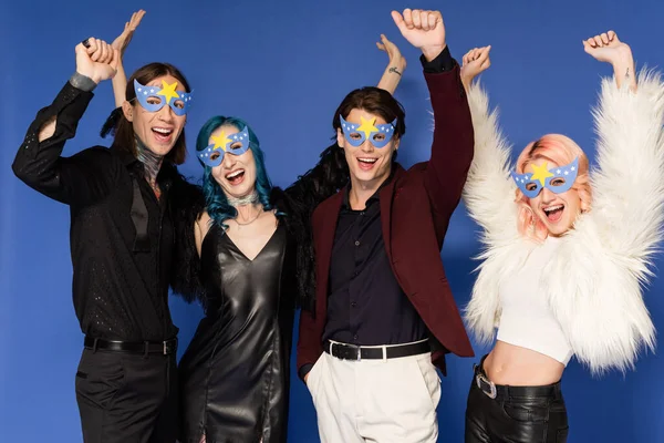 Joyful nonbinary friends in fashionable clothes and party masks standing with raised hands isolated on blue — Stock Photo