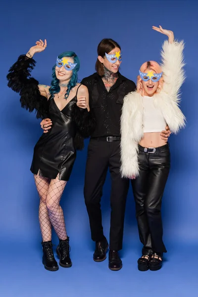 Full length of tattooed queer person hugging cheerful nonbinary friends in party masks waving hands on blue background — Stock Photo