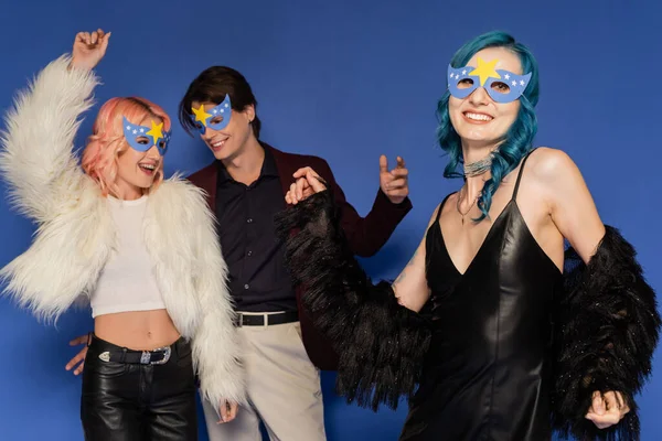 Cheerful queer people in faux fur jackets dancing near nonbinary friend at christmas party isolated on blue — Stock Photo