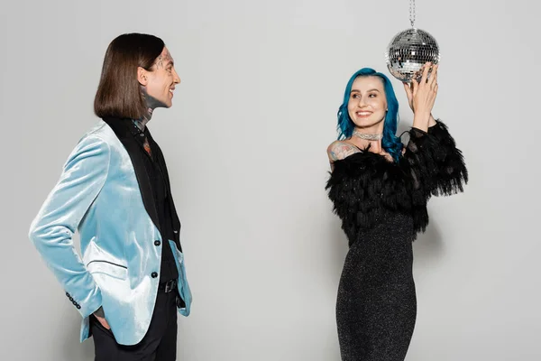 Smiling queer person in blue wig touching disco ball near elegant nonbinary friend on grey background — Stock Photo
