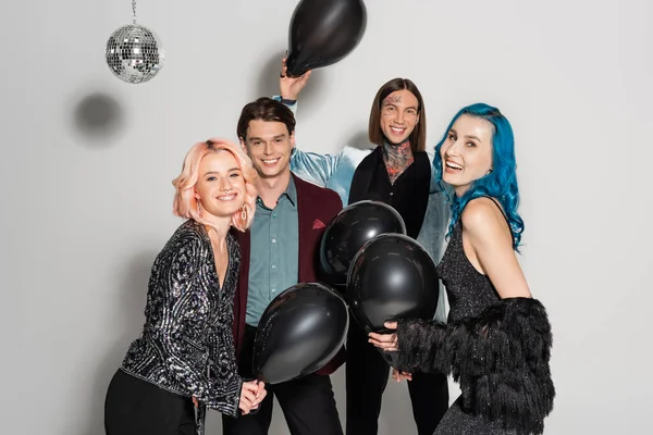 Erfreut queere Menschen mit schwarzen festlichen Luftballons, die vor grauem Hintergrund in die Kamera lächeln — Stockfoto
