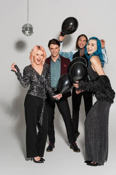 Full length of cheerful and stylish nonbinary friends with black balloons looking at camera on grey background — Stock Photo