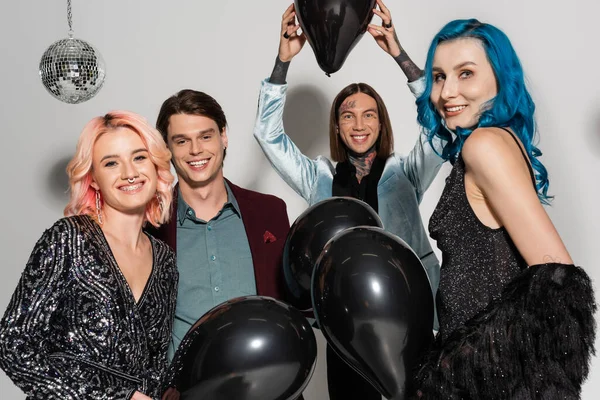 Cheerful queer person holding black balloon above head near nonbinary friends smiling at camera on grey background — Stock Photo