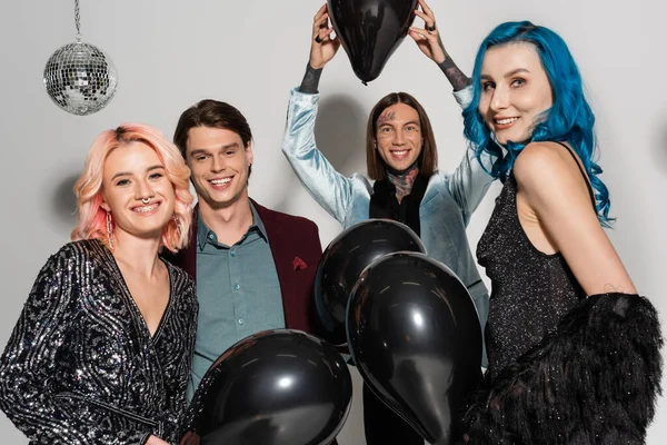 Alegre queer pessoa segurando preto balão acima da cabeça perto de amigos não binários sorrindo para câmera no fundo cinza — Fotografia de Stock