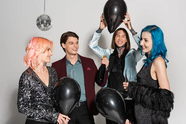 Fröhlich tätowierte queere Person mit schwarzem Luftballon über dem Kopf in der Nähe nichtbinärer Freunde, die auf grauem Hintergrund reden — Stockfoto