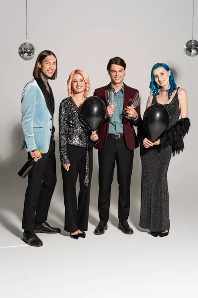 Pleine longueur de personnes queer heureux en vêtements de fête tenant champagne et ballons noirs sur fond gris — Photo de stock